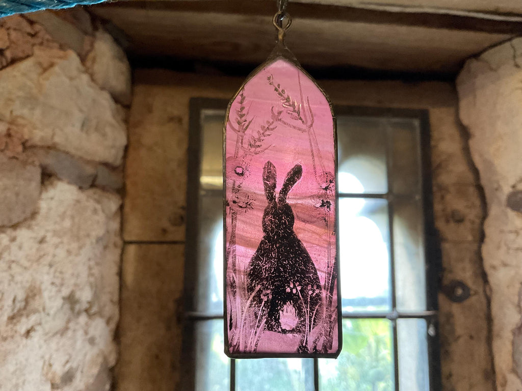 Decorative stained glass panel with a rabbit design hanging against a leaded window surrounded by a stone wall.
