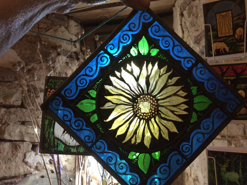 Decorative stained glass panel with floral sunflower design and blue swirly border, held against a window in an old stone wall.  The pieces of glass have been hand painted and kiln fired in the traditional manner