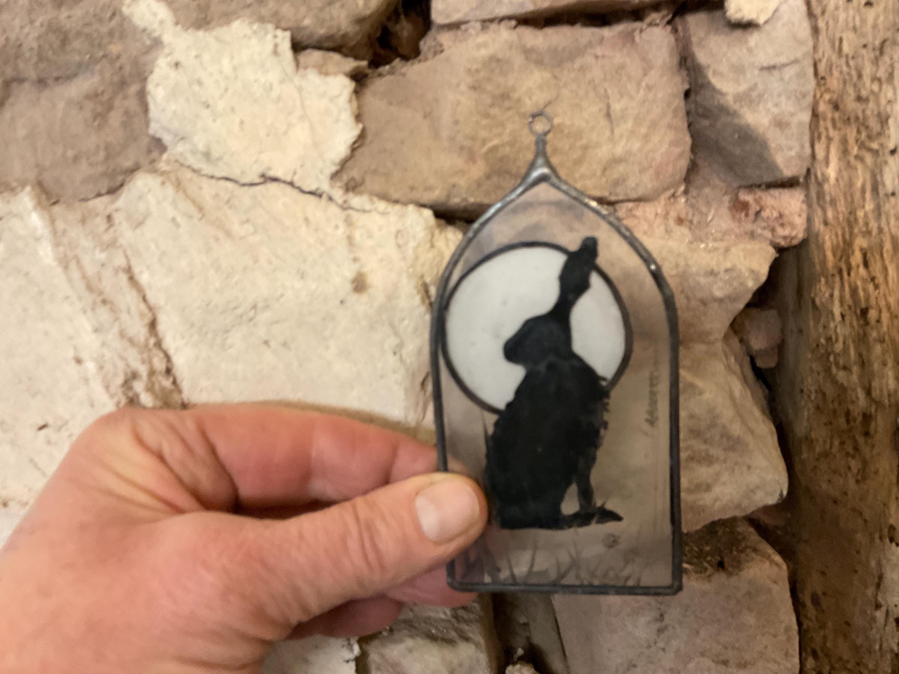 Hand holding a decorative glass panel with a black silhouette of a rabbit against a white circle, against a stone wall background.