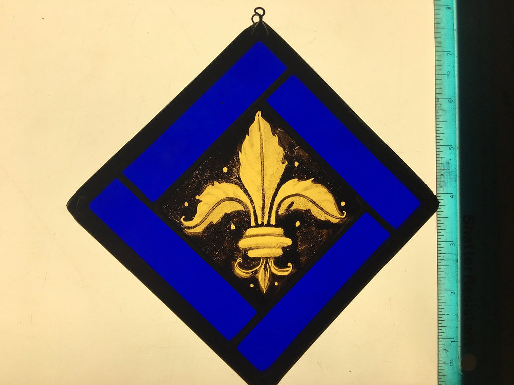 Decorative stained glass panel with gold fleur-de-lis design hand painted and kiln fired on a blue background, next to a ruler for scale.