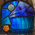 Stained glass panel with a magpie, moon, and pentacle design in blue, teal and black tones.