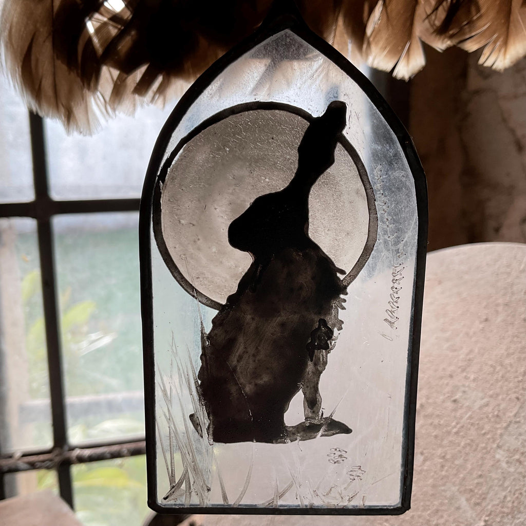 Decorative stained glass enamelled  panel with a silhouette of a hare or jack rabbit and moon, displayed by feathers in a window.