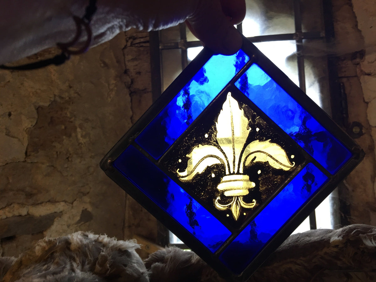 Stained glass panel with a gold fleur-de-lis design on a blue background, held against a stone wall.