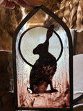 Stained glass decor of a silhouette hare in front of a full moon.  Held by hand with daylight and feathers