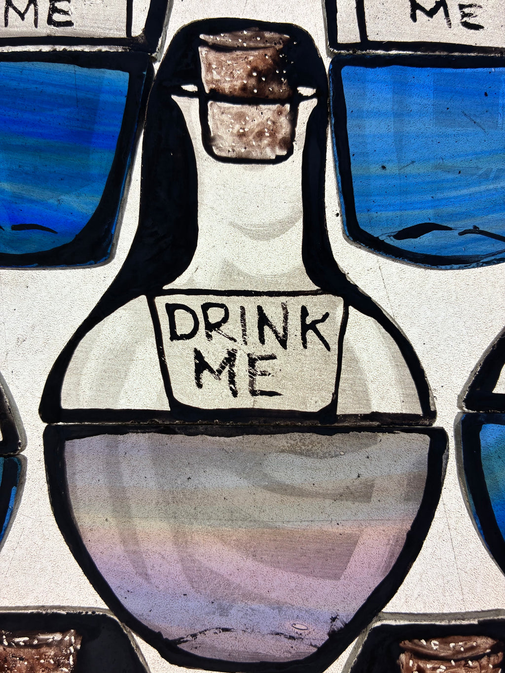 Hand painted stained glass depiction of a bottle labeled 'Drink Me' from a fantasy-themed design.