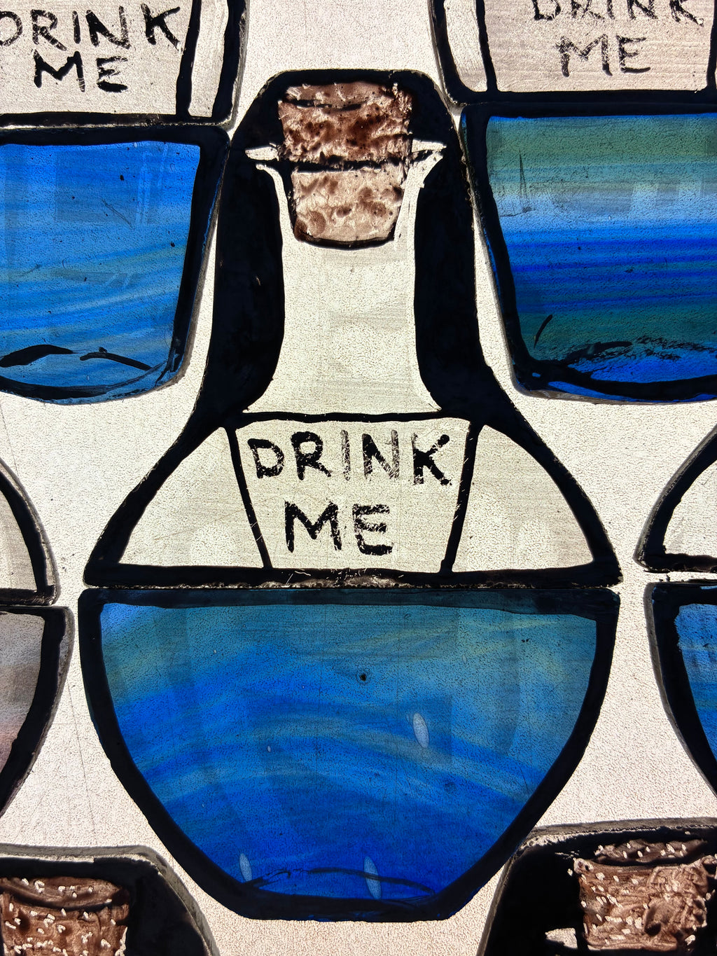 Blue potion bottle with 'Drink Me' label painted and kin fired in stained glass.  Shown on a light box as a “work in progress” image.