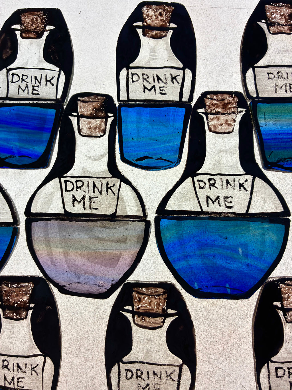 A collection of stained glass window decors inspire by Alice in Wonderland with 'Drink Me' painted on each of the bottles and blue liquid
