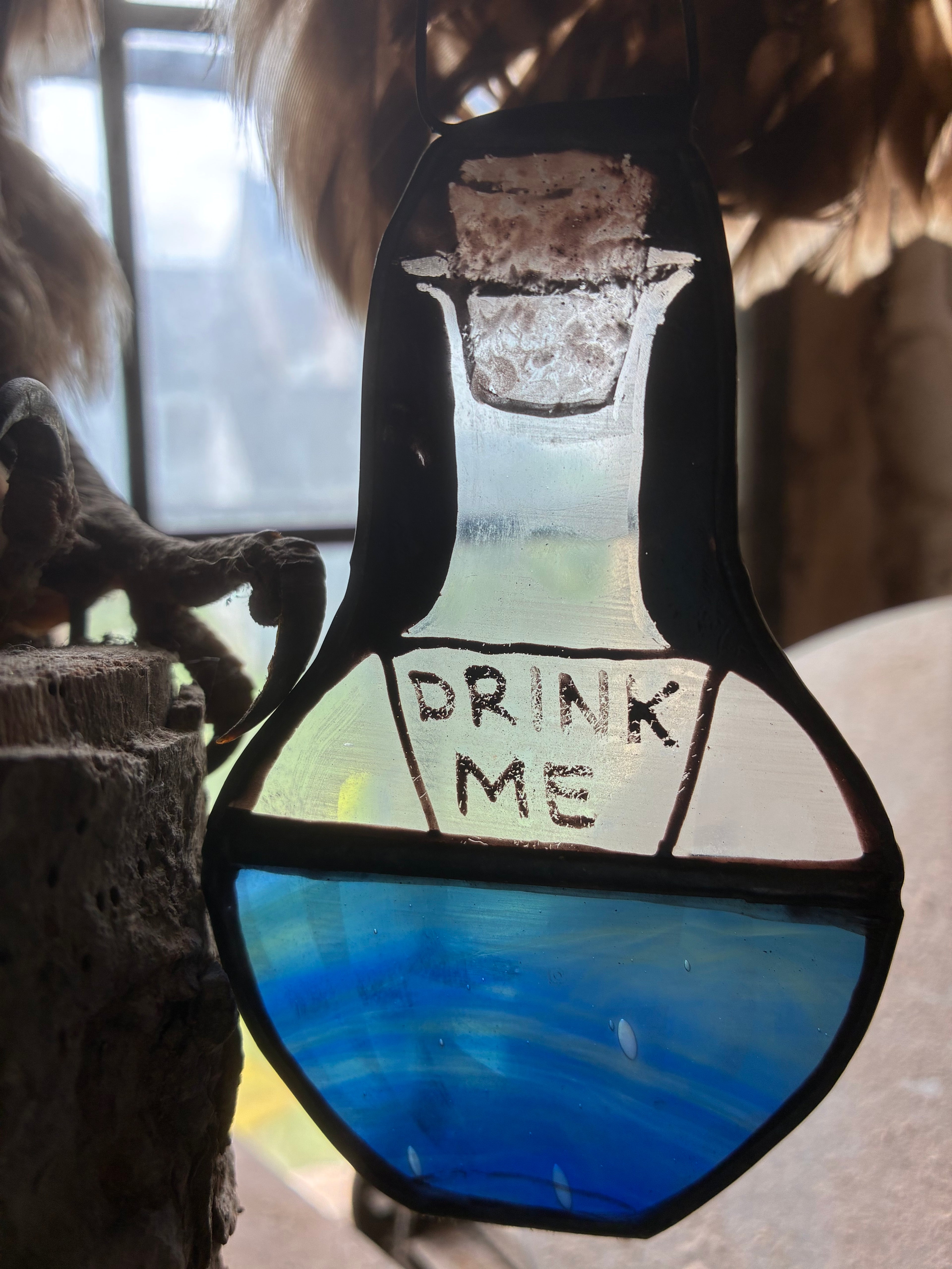 Sun catcher of a bottle with 'Drink Me' text on a blurred background with feathers and a claw