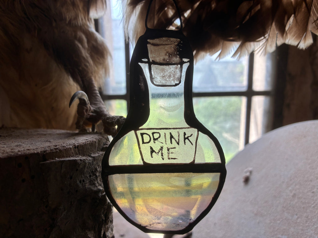 Glass stained glass bottle suncatcher with  DRINK ME text hand painted and kiln fired, shown with daylight and feathers.