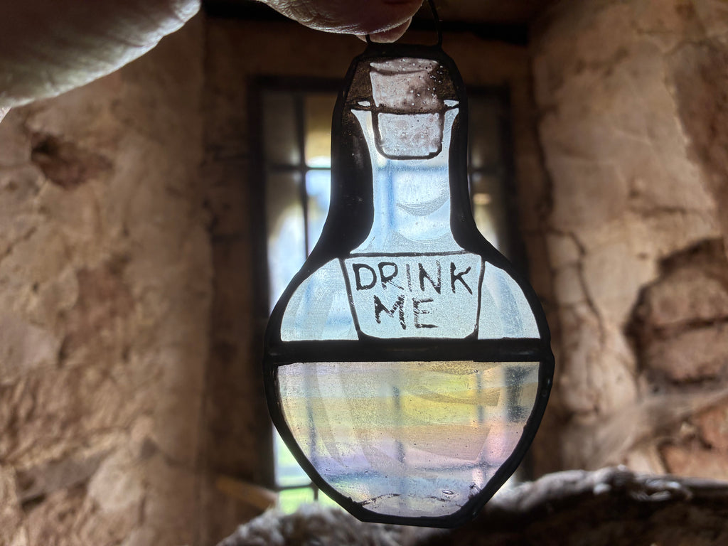 Stained glass light catcher with 'Drink Me' text against a stone wall background.