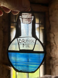 Hand painted and kiln fired stained glass bottle decoration with 'Drink Me' text held by a hand, blurred background.  Inspired by the story of Alice in Wonderland.