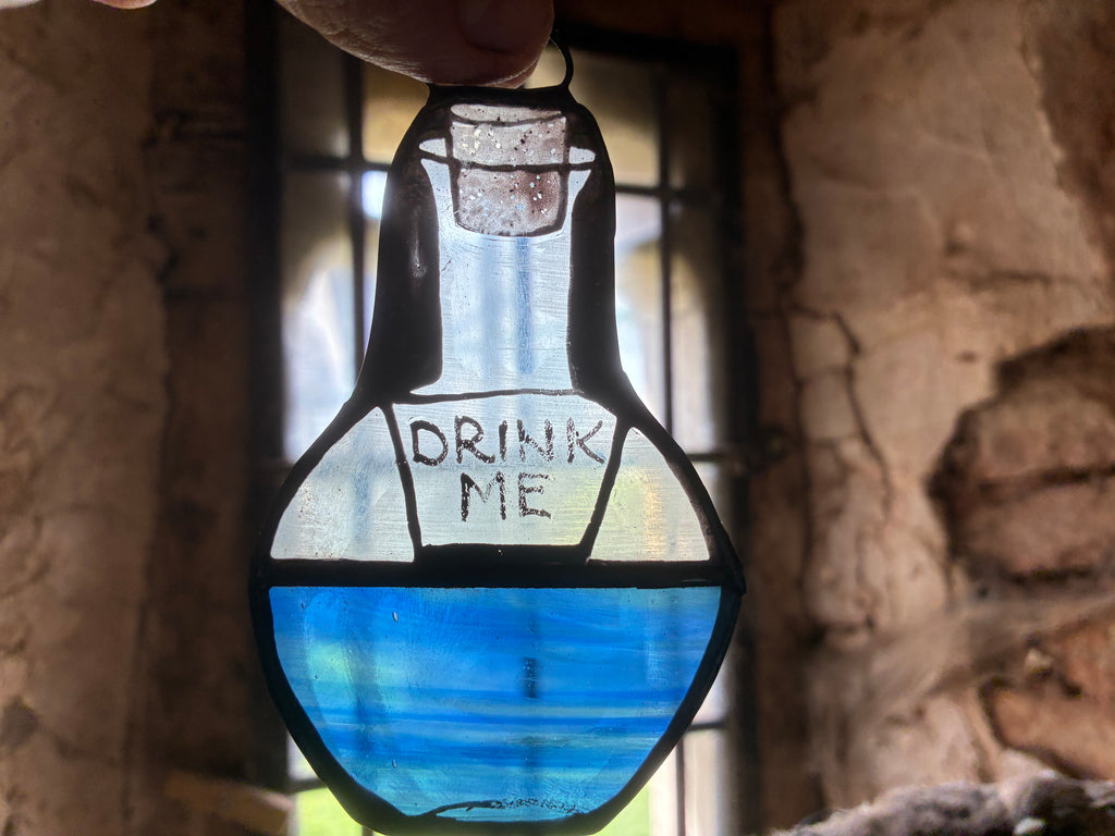 Stained glass pendant decor shaped like a bottle with blue liquid and 'DRINK ME' text, held against an old leaded window and a stone wall