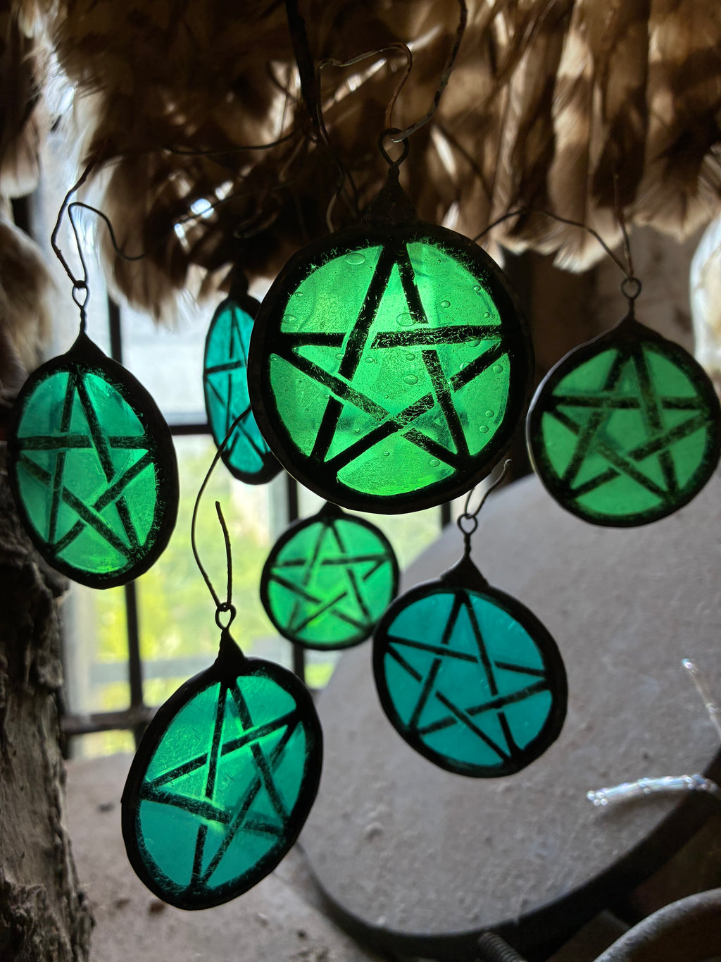 Green pentagram sun catchers hanging against a window with feathers.