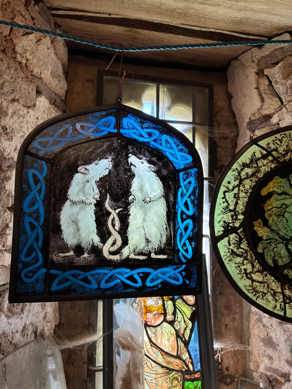 A stained glass window panel depicting two white rats on a black background, framed by blue Celtic knot borders, hanging in a rustic stone room.
