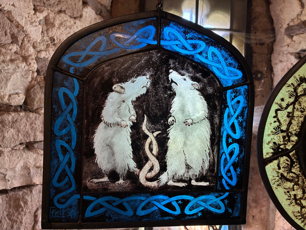 A stained glass window panel depicting two white rats facing each other with their tails entwined framed by bright blue Celtic knot borders, hanging against a rustic stone wall.