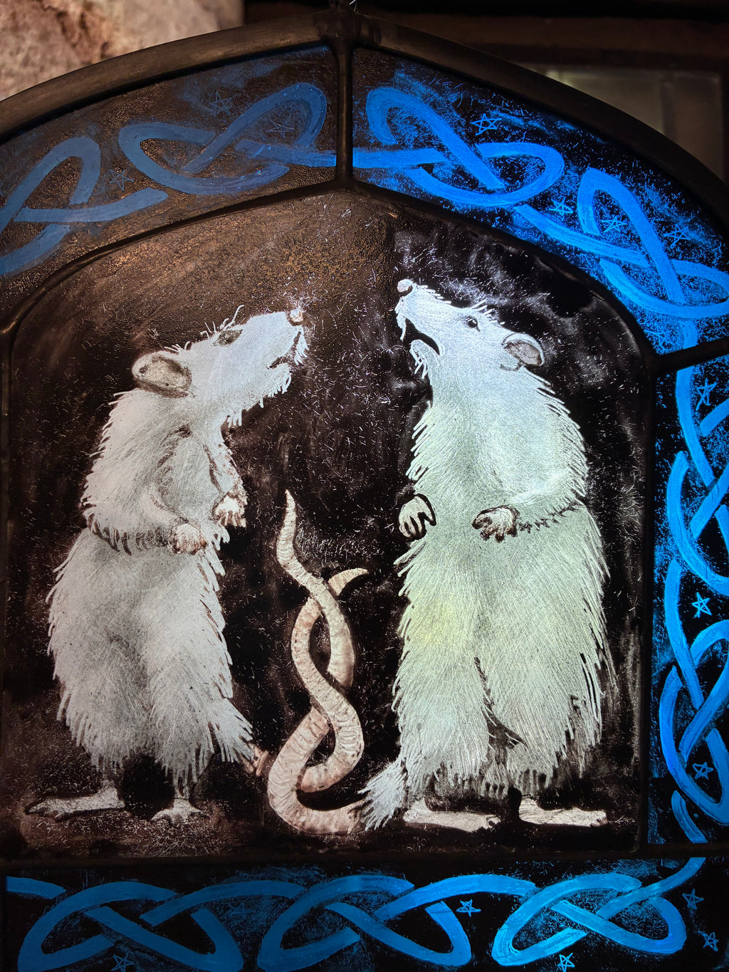 A stained glass window panel depicting two white rats facing each other with tails twisted together, framed by a bright blue Celtic knot border on a dark background.