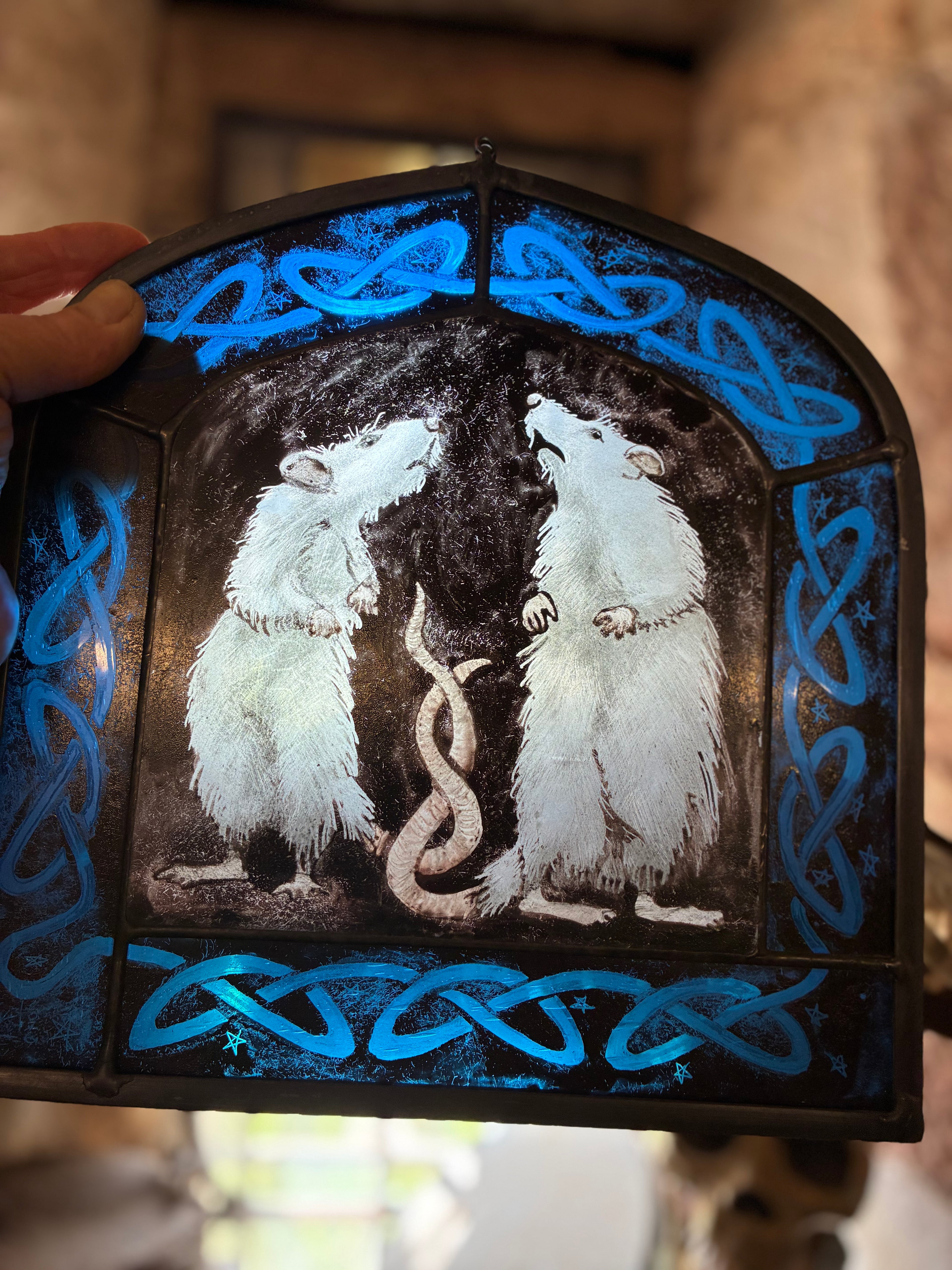 Stained glass panel showing two rats surrounded by blue glass with a Celtic knot design held by the hand that made it.