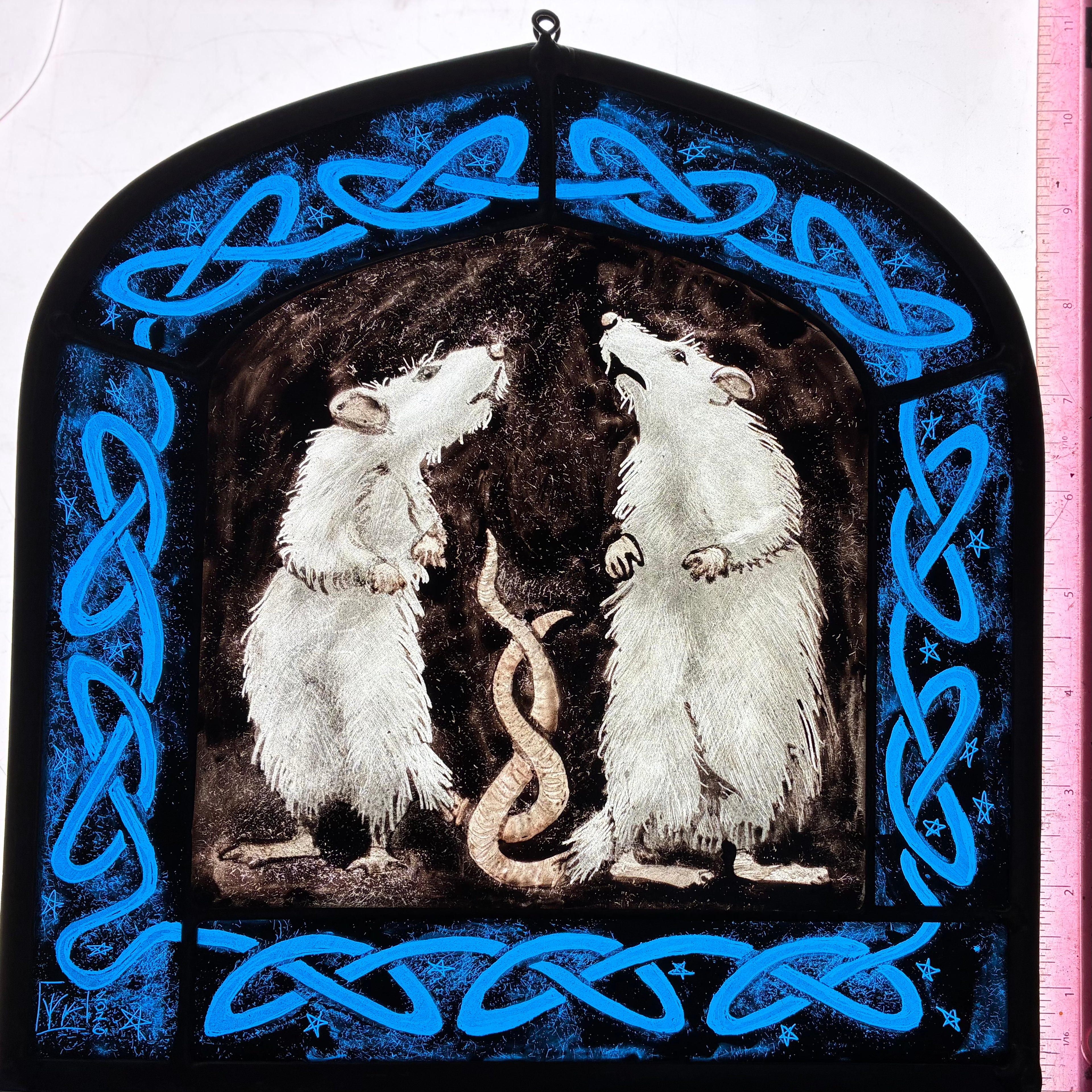 A stained glass window panel depicting two white rats facing each other with their tails entwined framed by a bright blue Celtic knot border on a black background.