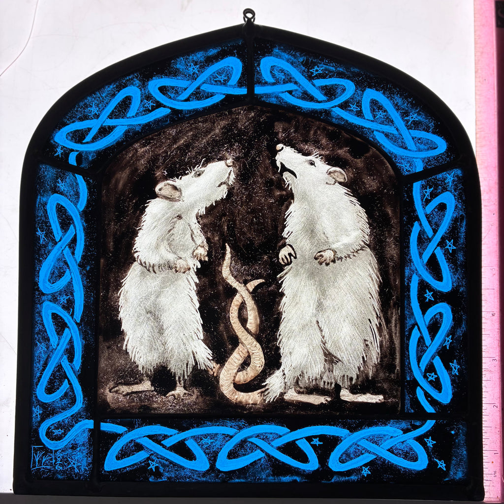 A stained glass window depicting two white rats facing each other, framed by a bright blue Celtic knot border on a black background, shown on a light box with a pink ruler for scale.