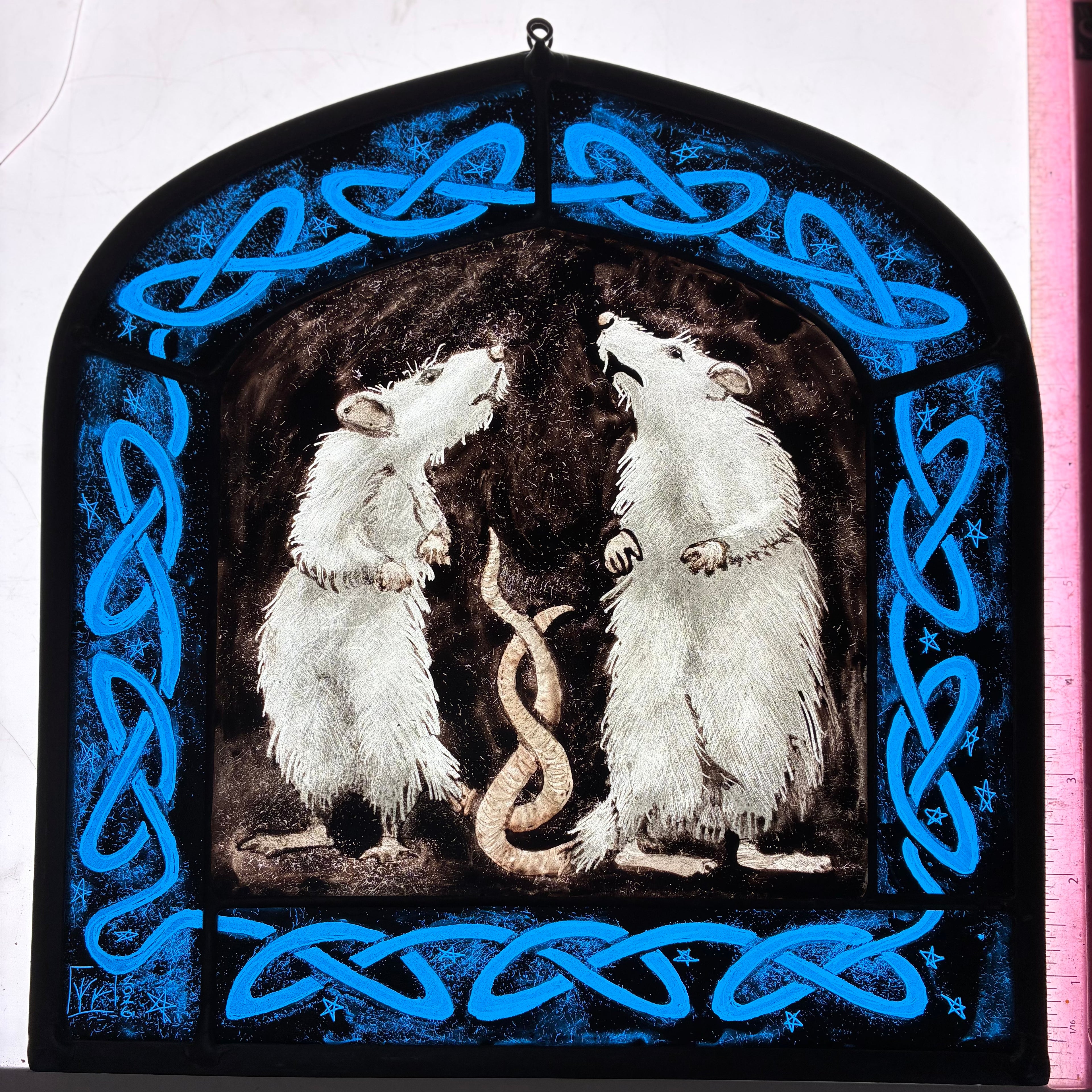 A stained glass window depicting two white rats facing each other, framed by a bright blue Celtic knot border on a black background, shown on a light box with a pink ruler for scale.