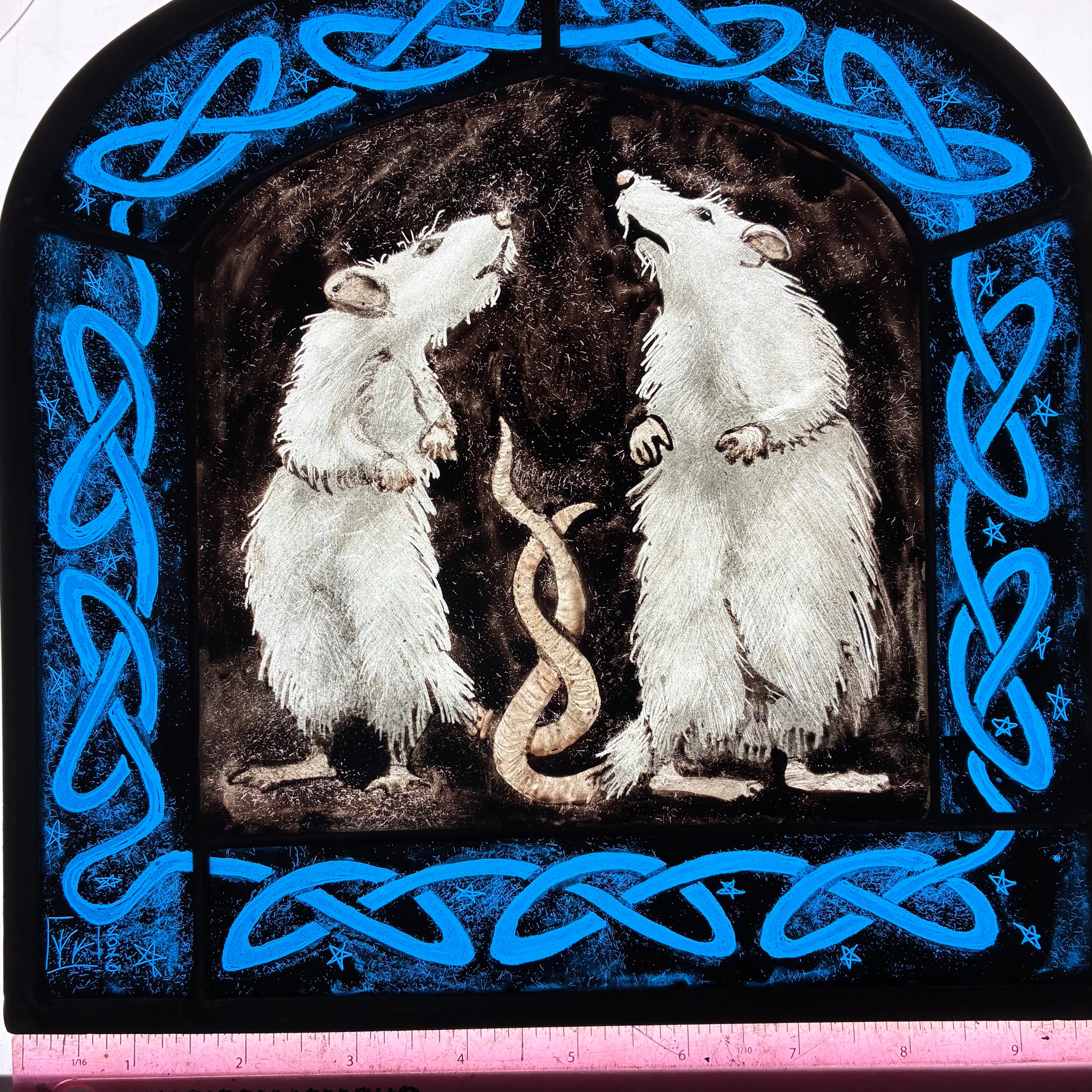 A square decorative stained glass artwork showing two white rats facing each other in a dark arched scene, framed by bright blue Celtic knot borders on a black background. A pink ruler is visible along the bottom edge for scale.