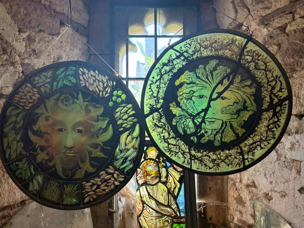 Two large circular stained glass green man panels hanging in a rustic stone interior. The left hand green man is for sale.  The panel features wheat, grass and leaf and berry motifs.