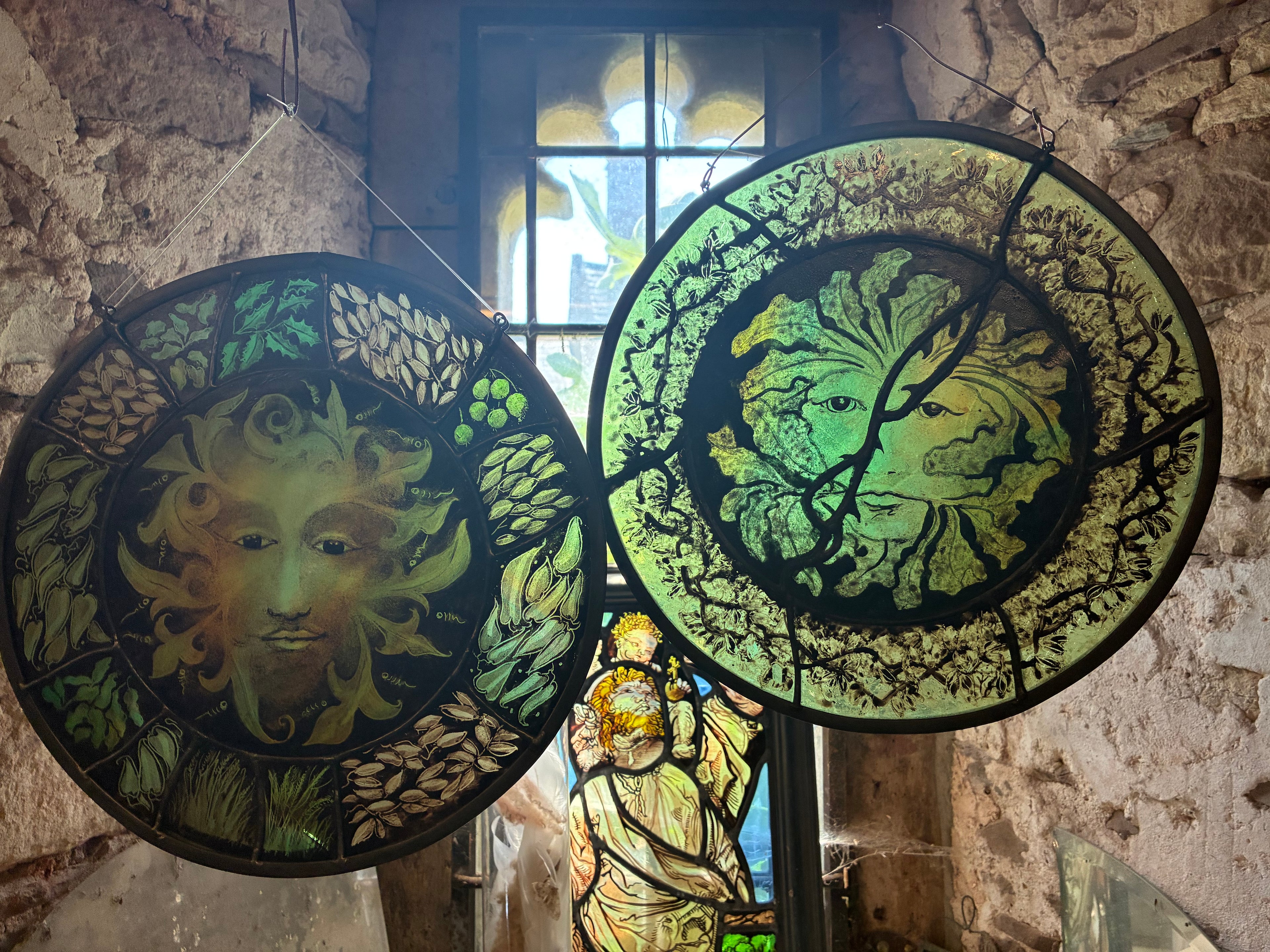 Two large circular stained glass green man panels hanging in a rustic stone interior. The left hand green man is for sale.  The panel features wheat, grass and leaf and berry motifs.