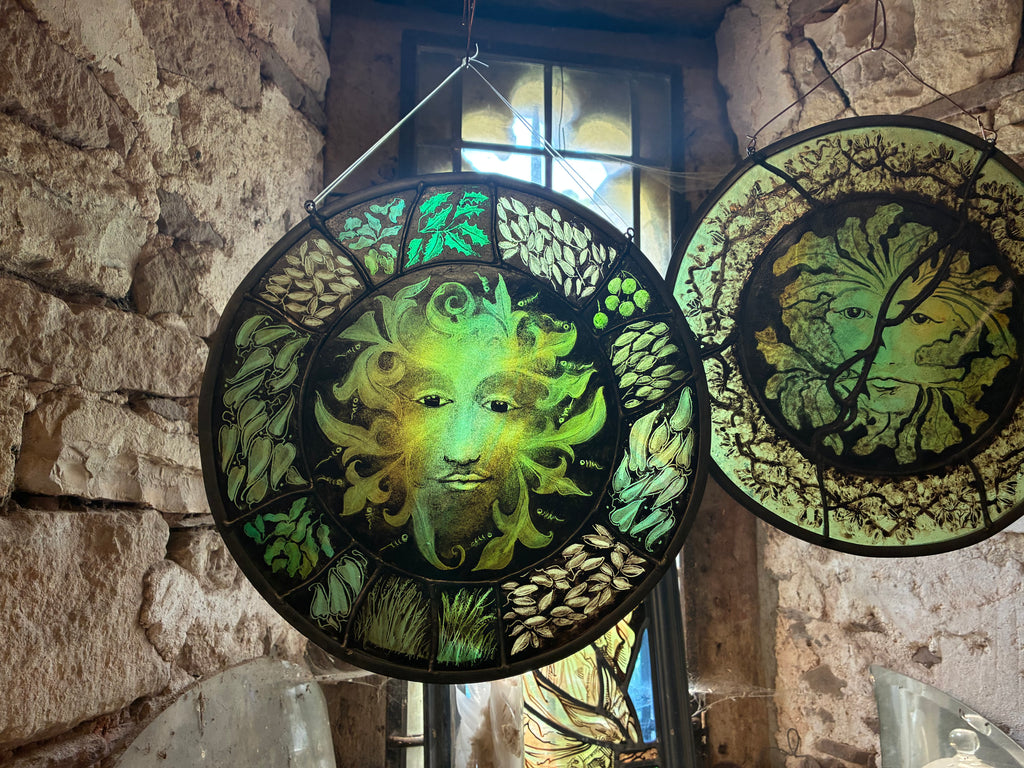 Two circular stained glass window panels featuring green and yellow tree-of-life and green man motifs, hanging in a rustic stone interior.  The left hand green man is for sale here.