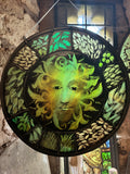 Green Man stained glass roundel, hand painted on hand blown glass, 12 inches, light shining through.  Green Man stained glass sun catcher roundel, hand painted and kiln fired shown hanging and backlit in front of an old window.