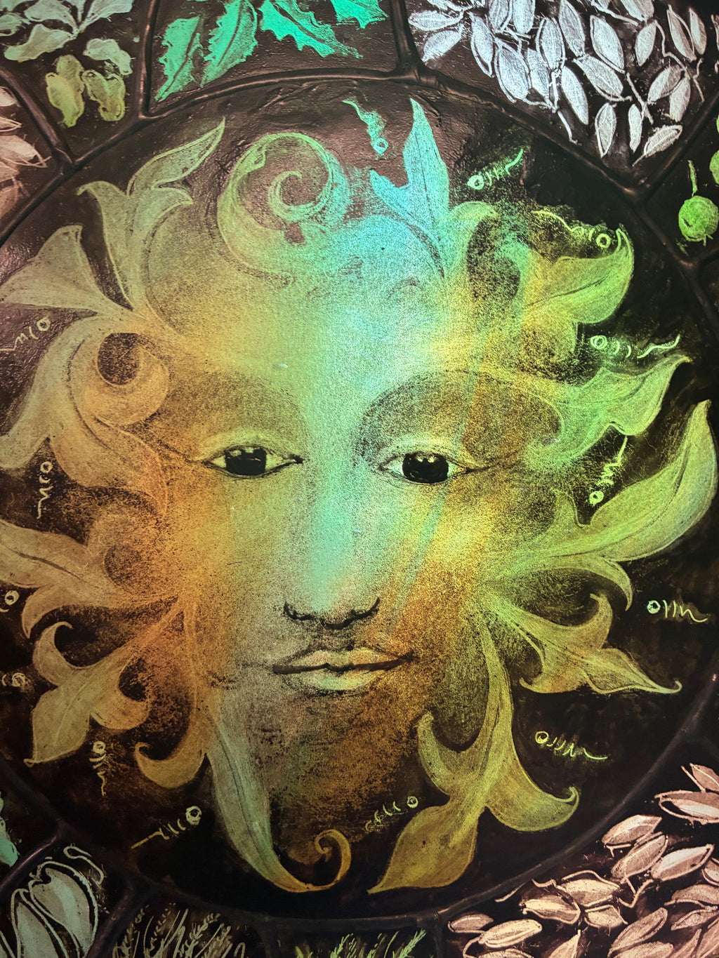 A close-up of a circular decorative artwork featuring a green man face with leaf-like hair in green, yellow, and gold tones on a dark background, surrounded by botanical motifs.