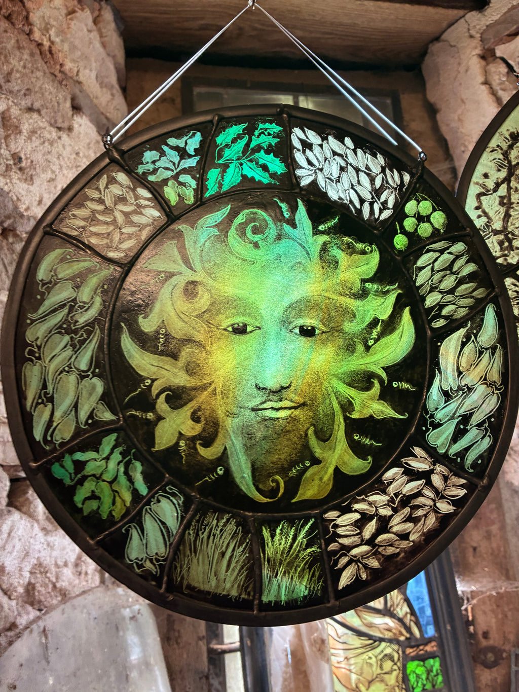 A circular stained glass window and hanging decorative panel featuring a central green  man  surrounded by leaf and vine motifs, set against a rustic stone interior background.