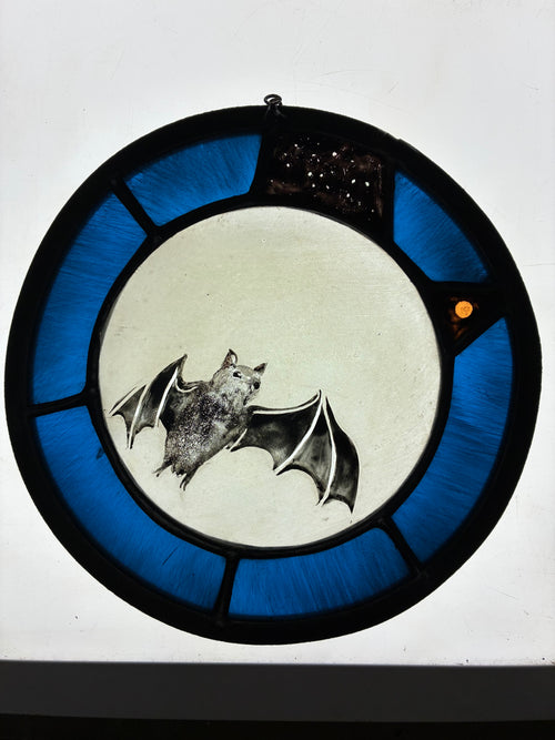 A circular stained glass window panel featuring a bat in front of a full moon, with a constellation and Mars, surrounded by bright blue sections and a black border, shown on a lit light box.