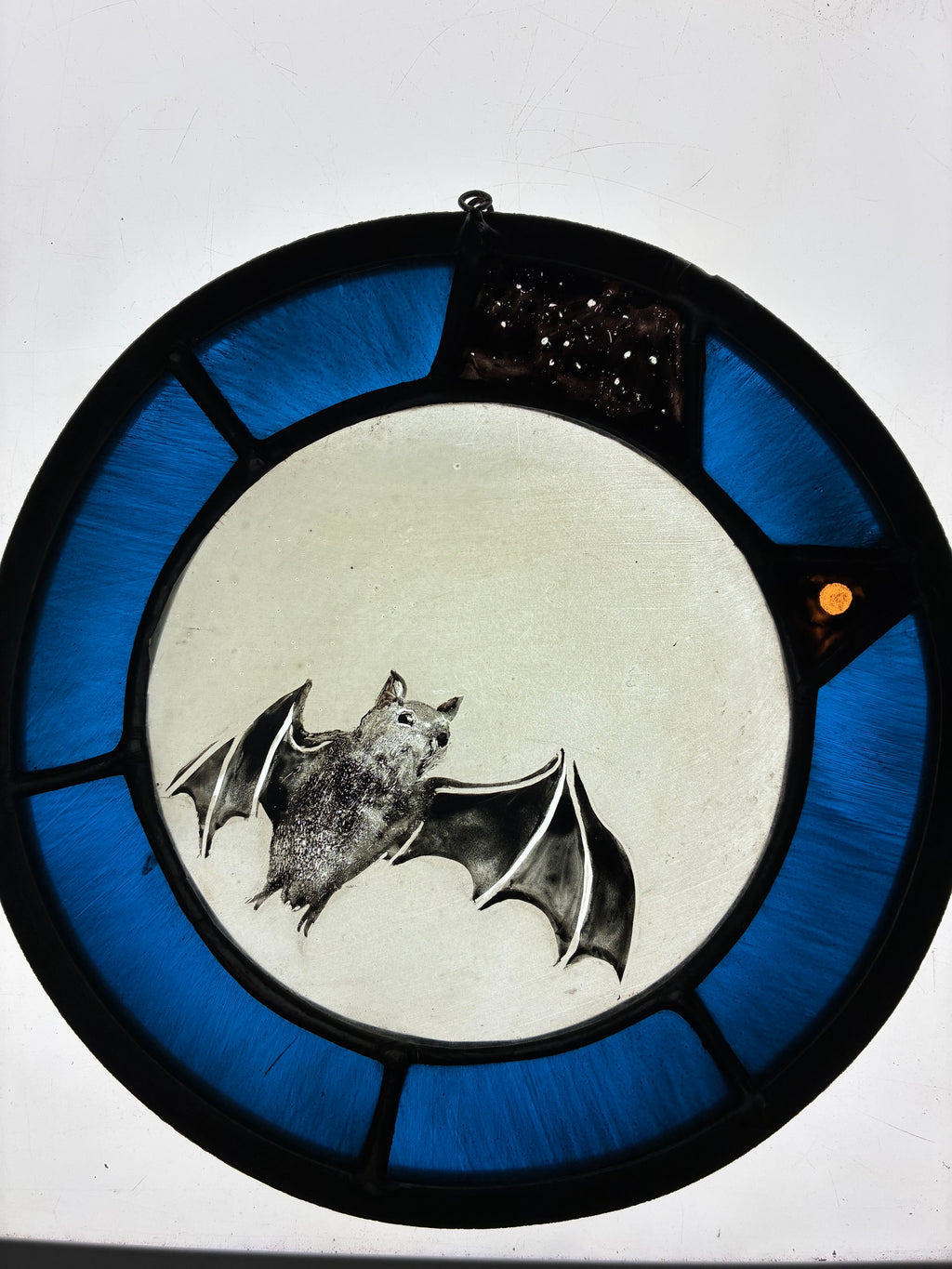 A circular stained glass window featuring a bat motif, with a white moon, a gray bat illustration, and a blue segmented border on a white background.