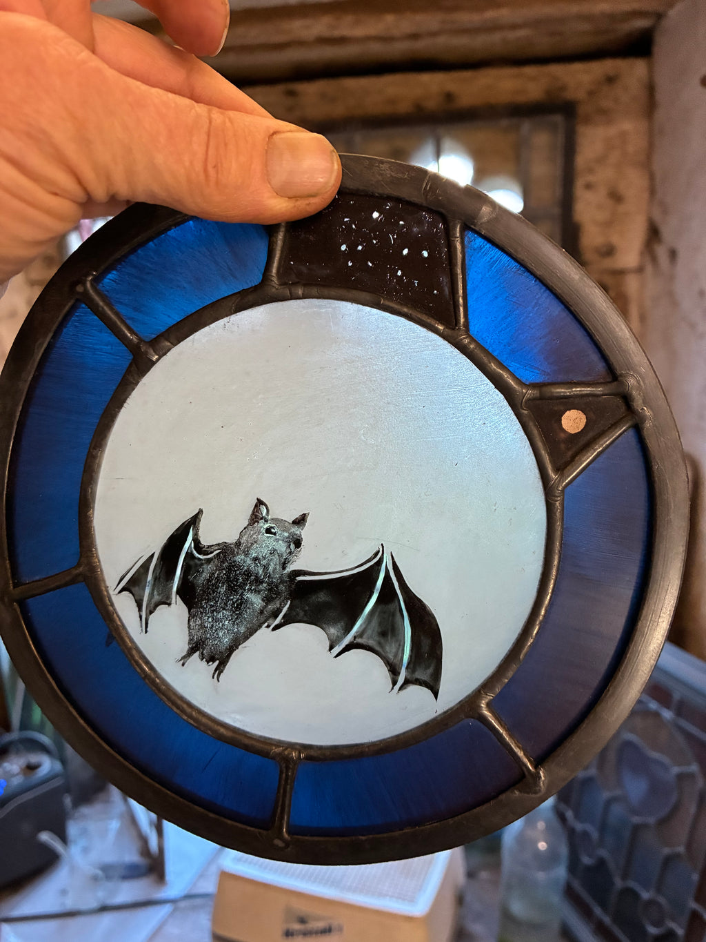 A hand holding a circular stained glass panel featuring a bat design, with blue glass sections and a dark metal frame.