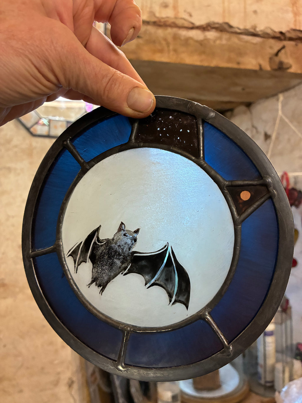 A hand holds a circular stained glass window featuring a bat design. The glass has a dark blue outer ring, a light blue-white moon center, and a black bat illustration with dark leaded metal borders.