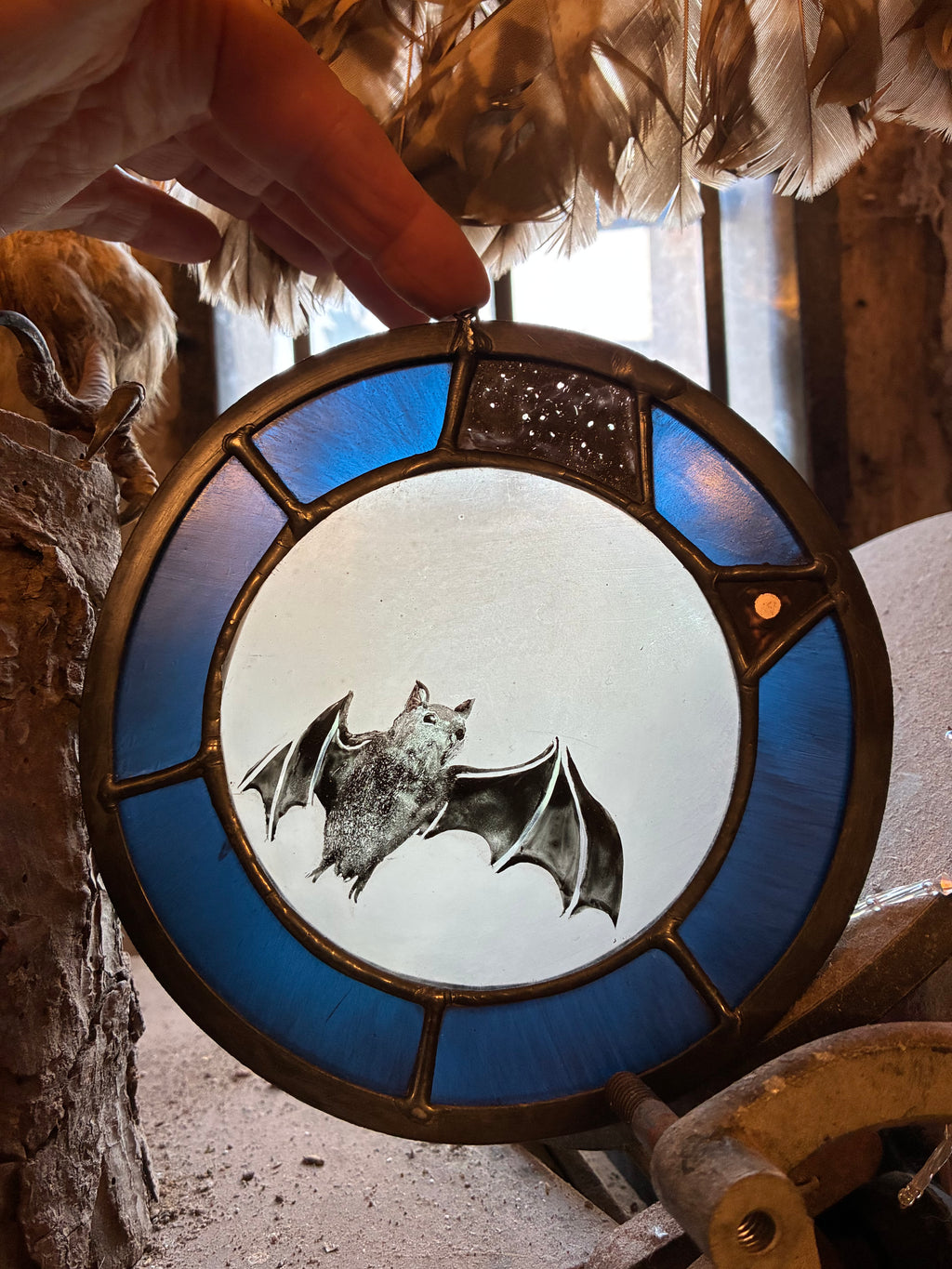 A hand holds a circular stained glass window panel featuring a bat design, with blue glass sections and a dark metal frame, set against a rustic background with feathers and wood.