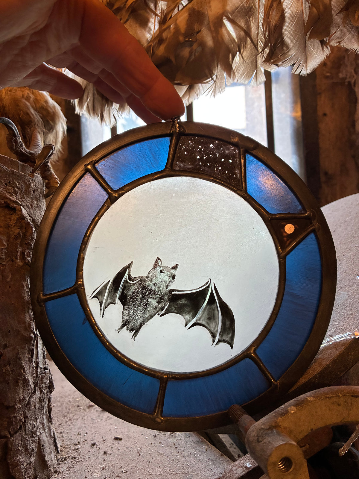 A hand holds a circular stained glass window panel featuring a bat design, with blue glass sections and a dark metal frame, set against a rustic background with feathers and wood.