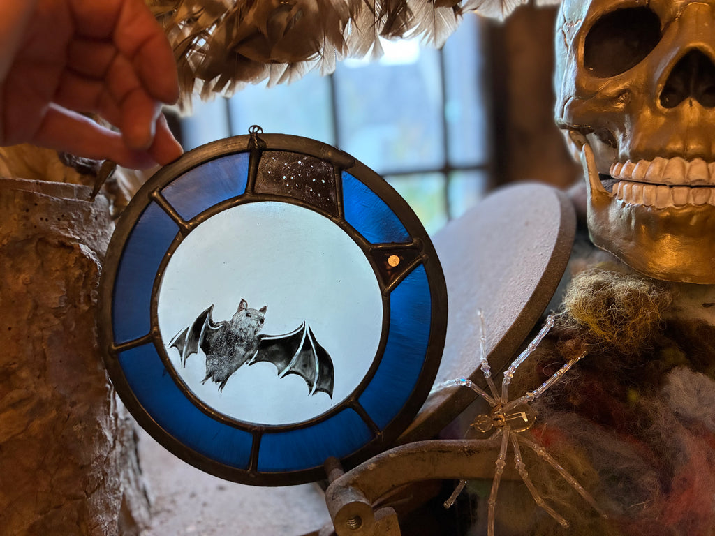 A circular stained glass window ornament featuring a bat design, held by a hand in a rustic Halloween-themed setting with a gold skull and spider nearby.