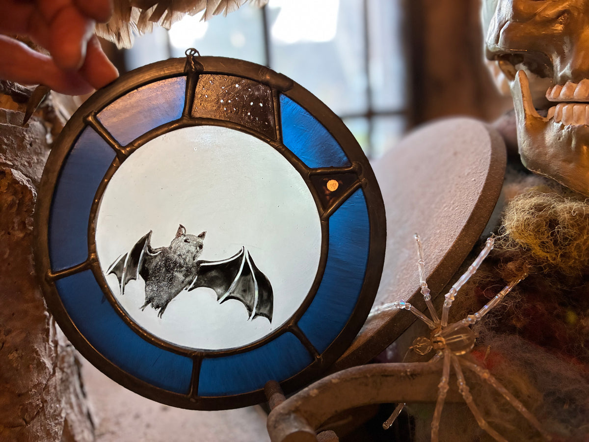 A circular stained glass hanging ornament featuring a bat design, held by a hand in a rustic, Halloween-themed setting with a skull and spider nearby.