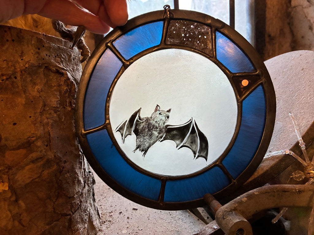 A circular stained glass suncatcher featuring a bat design, with the moon, a constellation and mars, held by a hand in front of a rustic stone background.