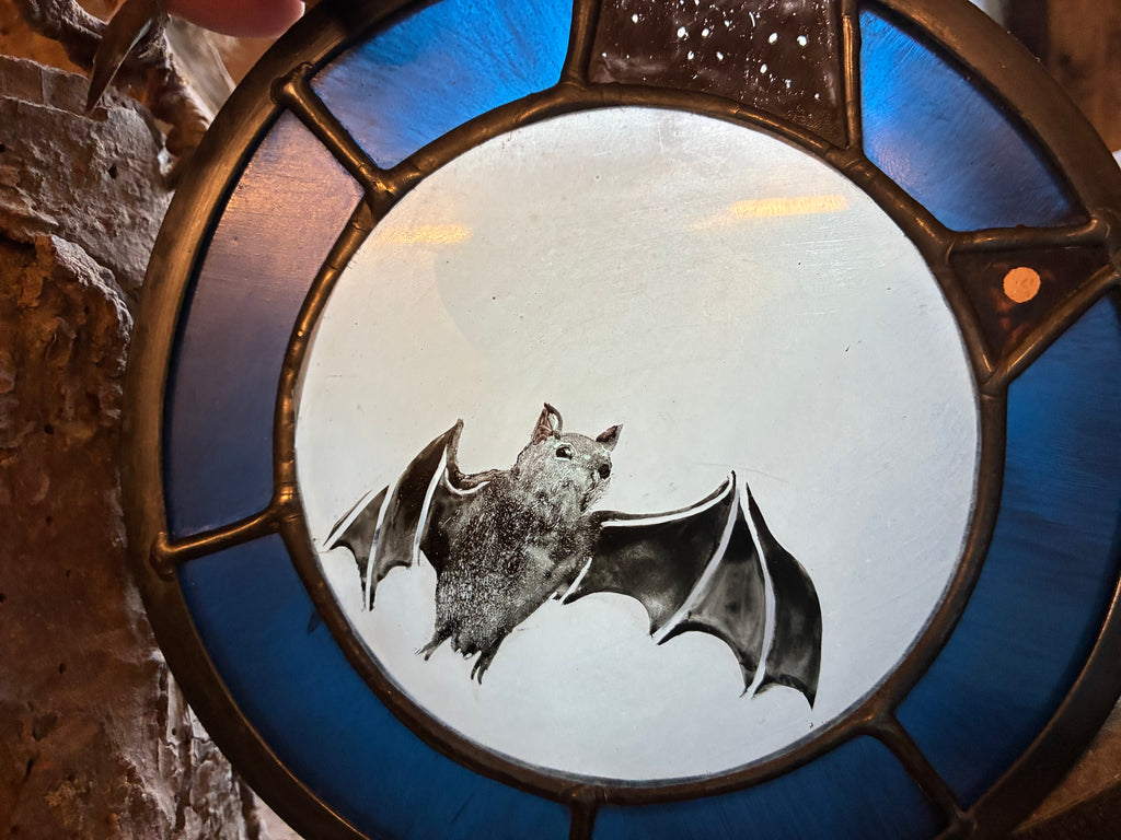 A circular stained glass window featuring a detailed bat illustration in the center, surrounded by blue glass panels and a dark metal frame, set against a rustic stone wall.