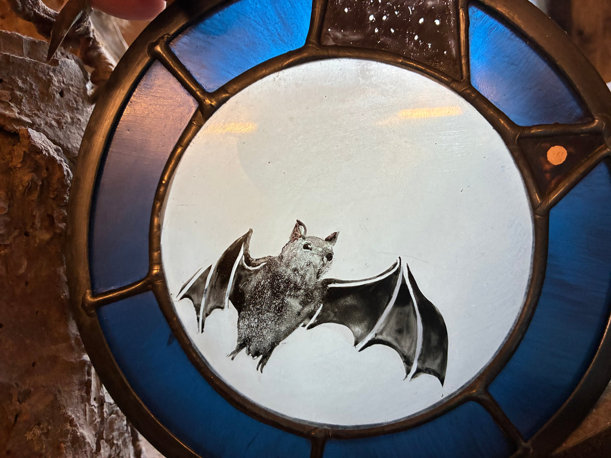 A circular stained glass window featuring a detailed bat illustration in the center, surrounded by blue glass panels and a dark metal frame, set against a rustic stone wall.