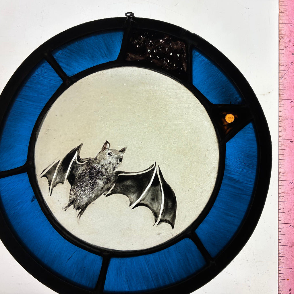 A circular stained glass window piece featuring a bat in the centre, flying axross the full moon and a blue outer ring intimating the night sly, with black leading and a small starry segment near the top. A pink measuring tape is visible along the right edge.