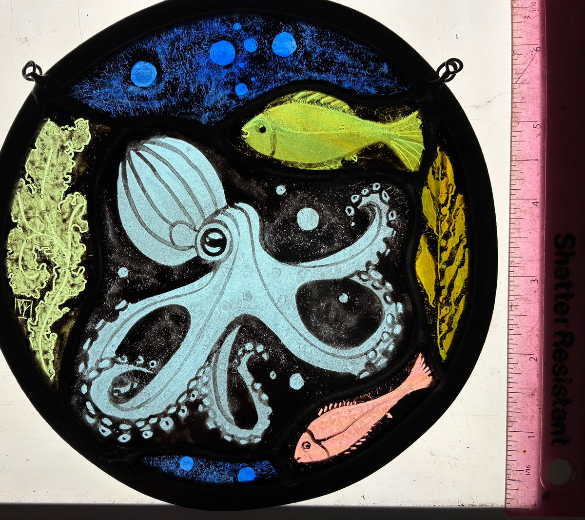 A circular stained glass artwork depicting an octopus, fish, seaweed, coral, and bubbles in blue, yellow, pink, and light blue tones on a black background, photographed on a light box with a pink ruler along the right edge.