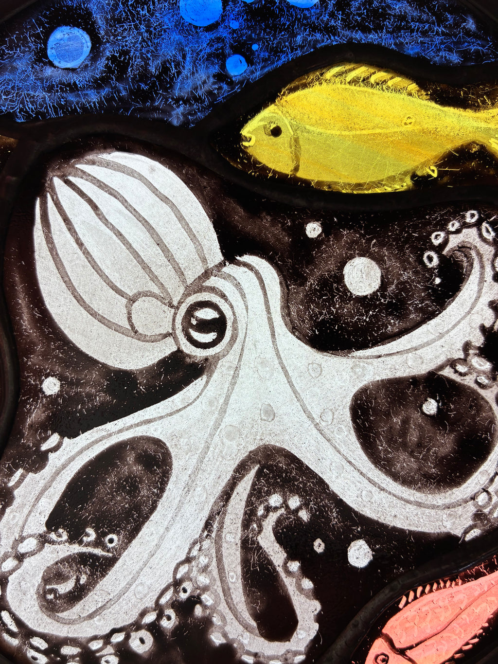 A close up of the  underwater-style artwork featuring a blue octopus on a black background, a bright green yellow fish near the top right, blue bubbles at the top, and a coral-pink fish at the bottom right.