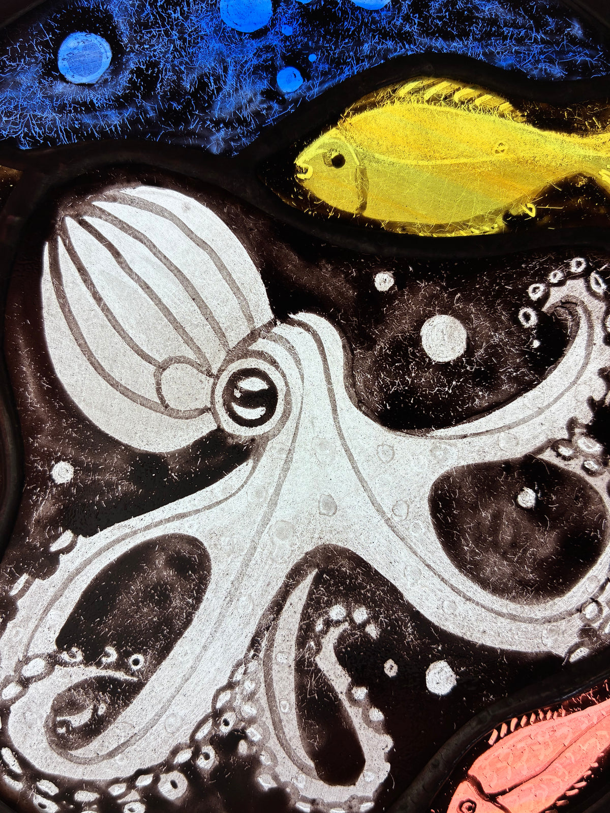 A close up of the  underwater-style artwork featuring a blue octopus on a black background, a bright green yellow fish near the top right, blue bubbles at the top, and a coral-pink fish at the bottom right.