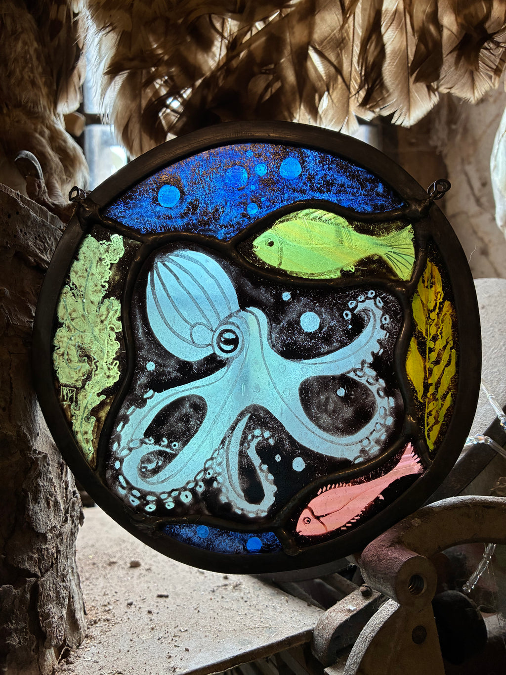 A round stained glass window featuring an underwater scene with a light blue octopus, a green fish, a pink fish, and blue bubbles on a black background, suspended in a rustic setting with feathers, wood and metal.
