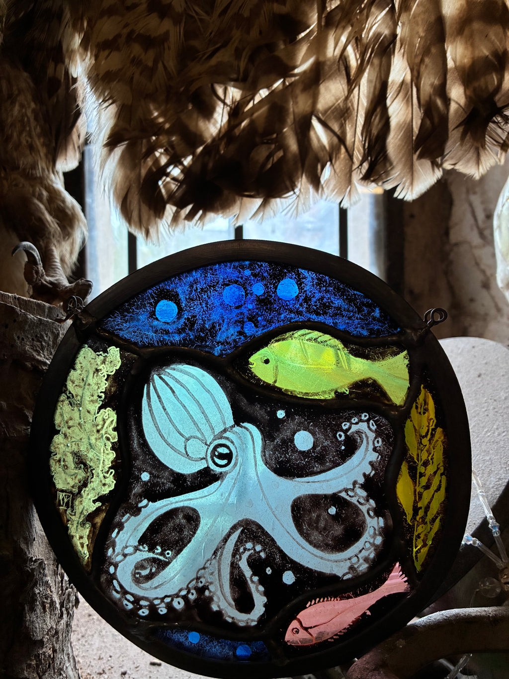 A circular stained glass window featuring an underwater scene with a light blue octopus, a bright yellow-green fish, a pink fish, and blue sea life on a black background, hanging in front of a window with fur and wood elements.