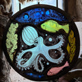 A circular stained glass art piece depicting an underwater scene with a large pale blue octopus, a bright green fish, a pink fish, green coral, and a dark blue background, framed in a dark metal hoop.