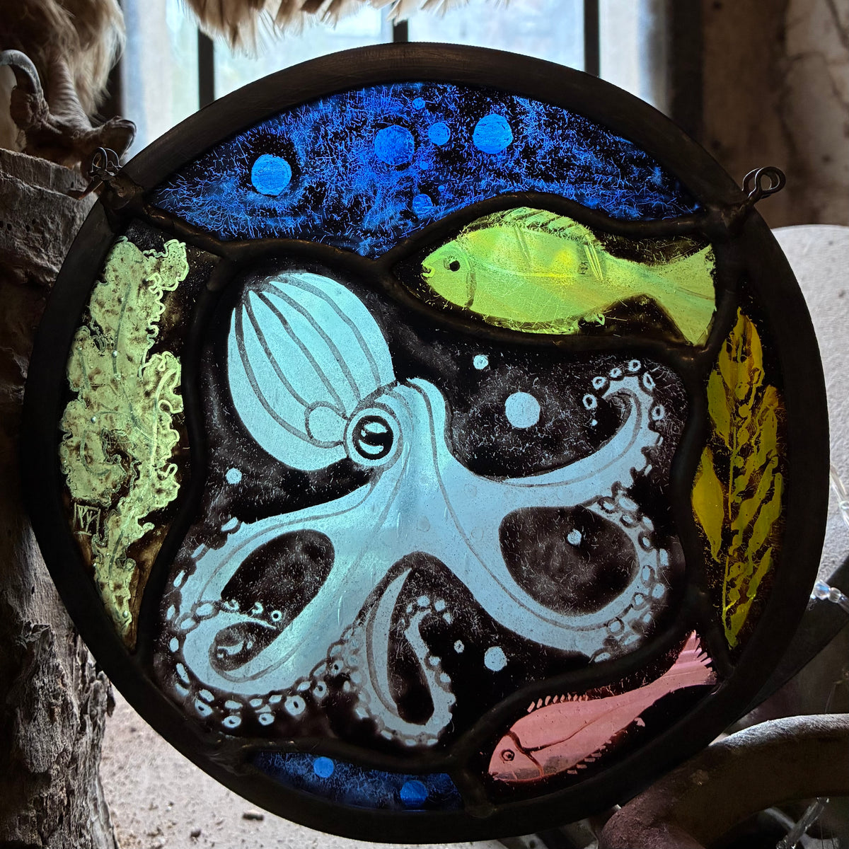 A circular stained glass art piece depicting an underwater scene with a large pale blue octopus, a bright green fish, a pink fish, green coral, and a dark blue background, framed in a dark metal hoop.